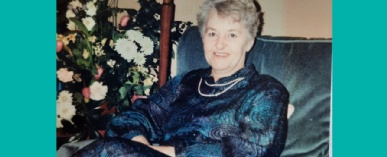 Daphne is dressed in a blue dress and pearls, smiling at the camera, seated on a blue velvet sofa with a large bouquet of flowers next to it.